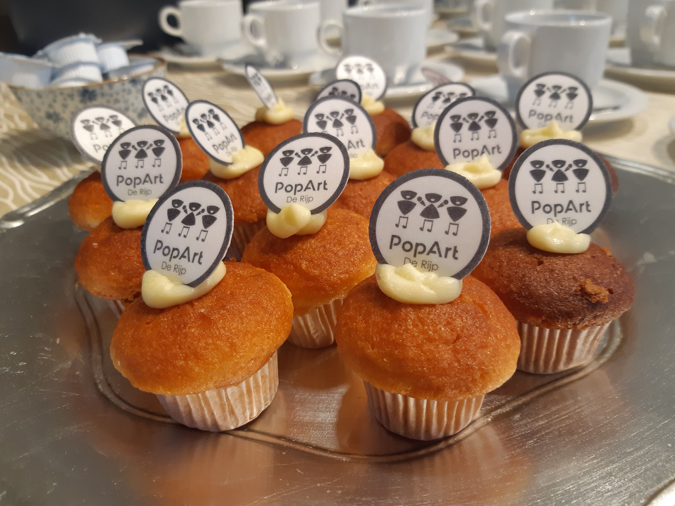 Goudbruine muffins met een beetje witte botercreme erop met daarin heen rondje gestoken waar het nieuwe PopArt logo op staat. 
